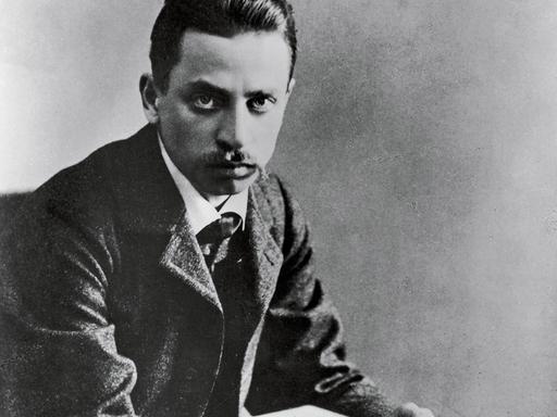 Schwarzweißporträt von Rainer Maria Rilke, der von einem aufgeschlagenen Buch aufblickt. Schwarzweißporträt von Rainer Maria Rilke, der von einem aufgeschlagenen Buch aufblickt.