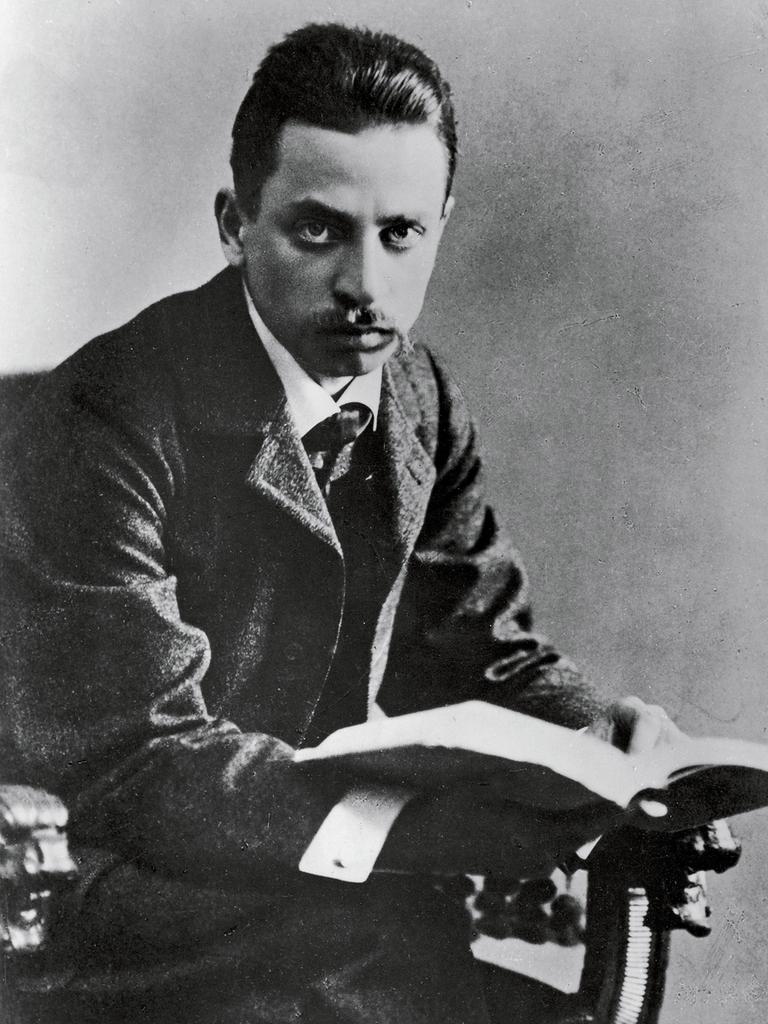 Schwarzweißporträt von Rainer Maria Rilke, der von einem aufgeschlagenen Buch aufblickt. Schwarzweißporträt von Rainer Maria Rilke, der von einem aufgeschlagenen Buch aufblickt.