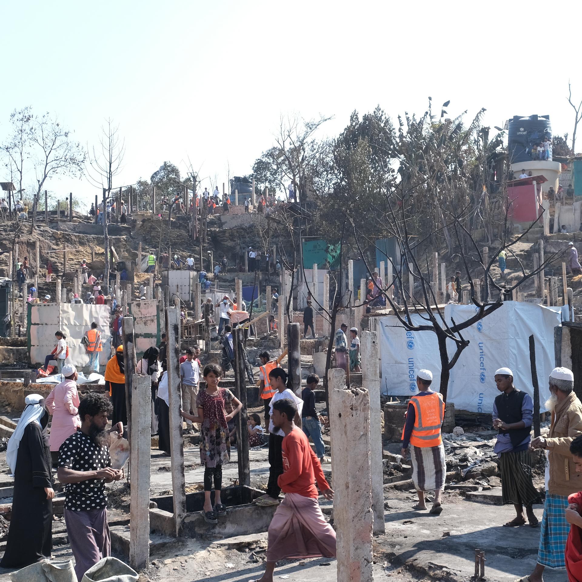 Menschen stehen im Lager zwischen zerstörten Zelten und Hütten.