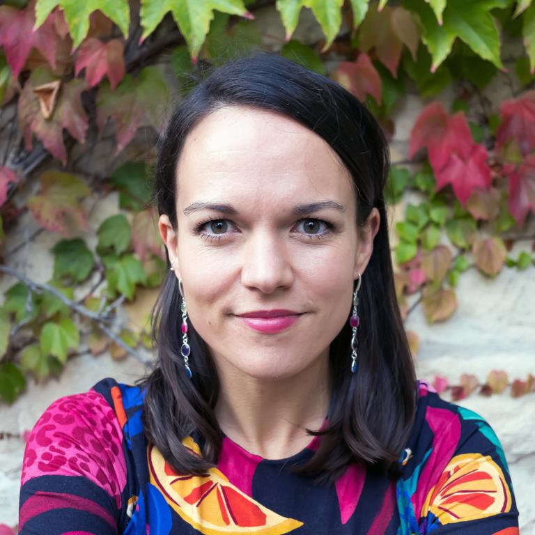 Porträt von Amrei Bahr in einer Bluse mit buntem Muster, mit grünem und rotem Weinlaub im Hintergrund