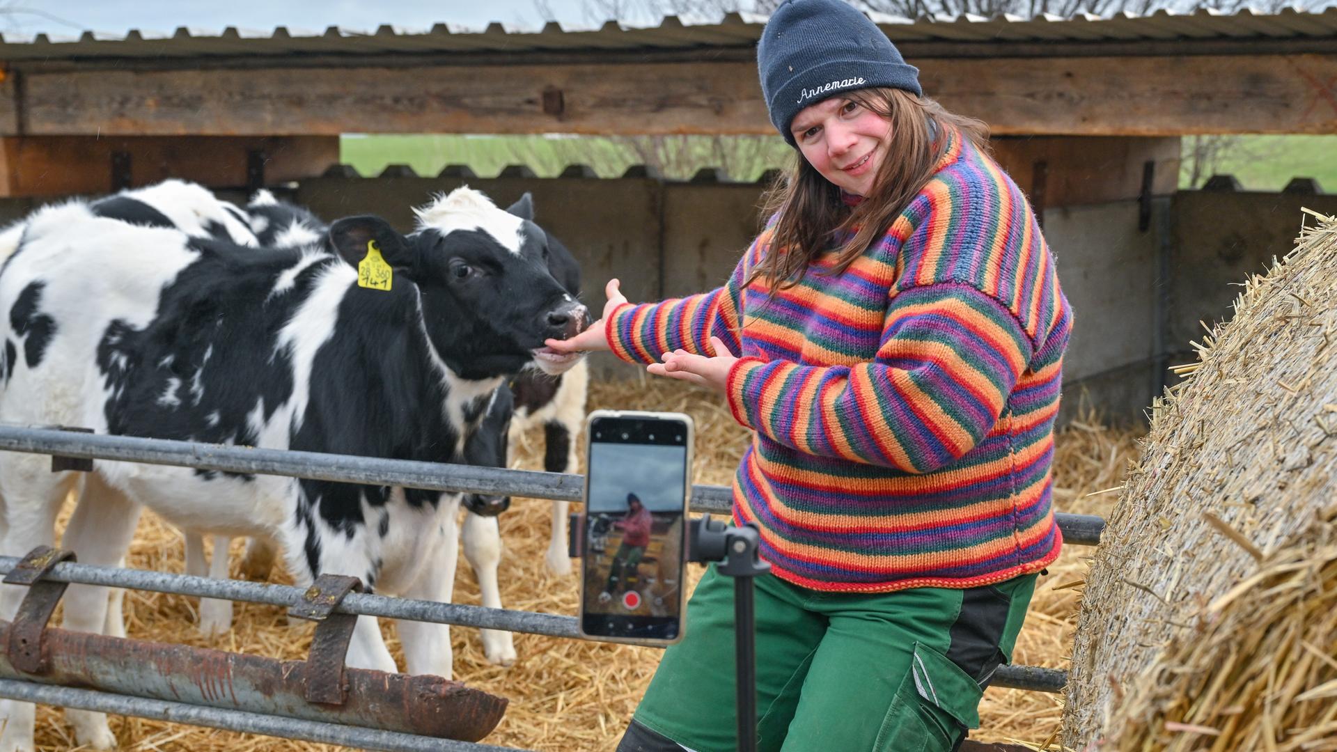 Eine junge Bäuerin steht in Gummistiefeln neben einer Kuh. Im Vordergrund Heuballen und ein Handy auf Stativ, das das Szenario filmt.