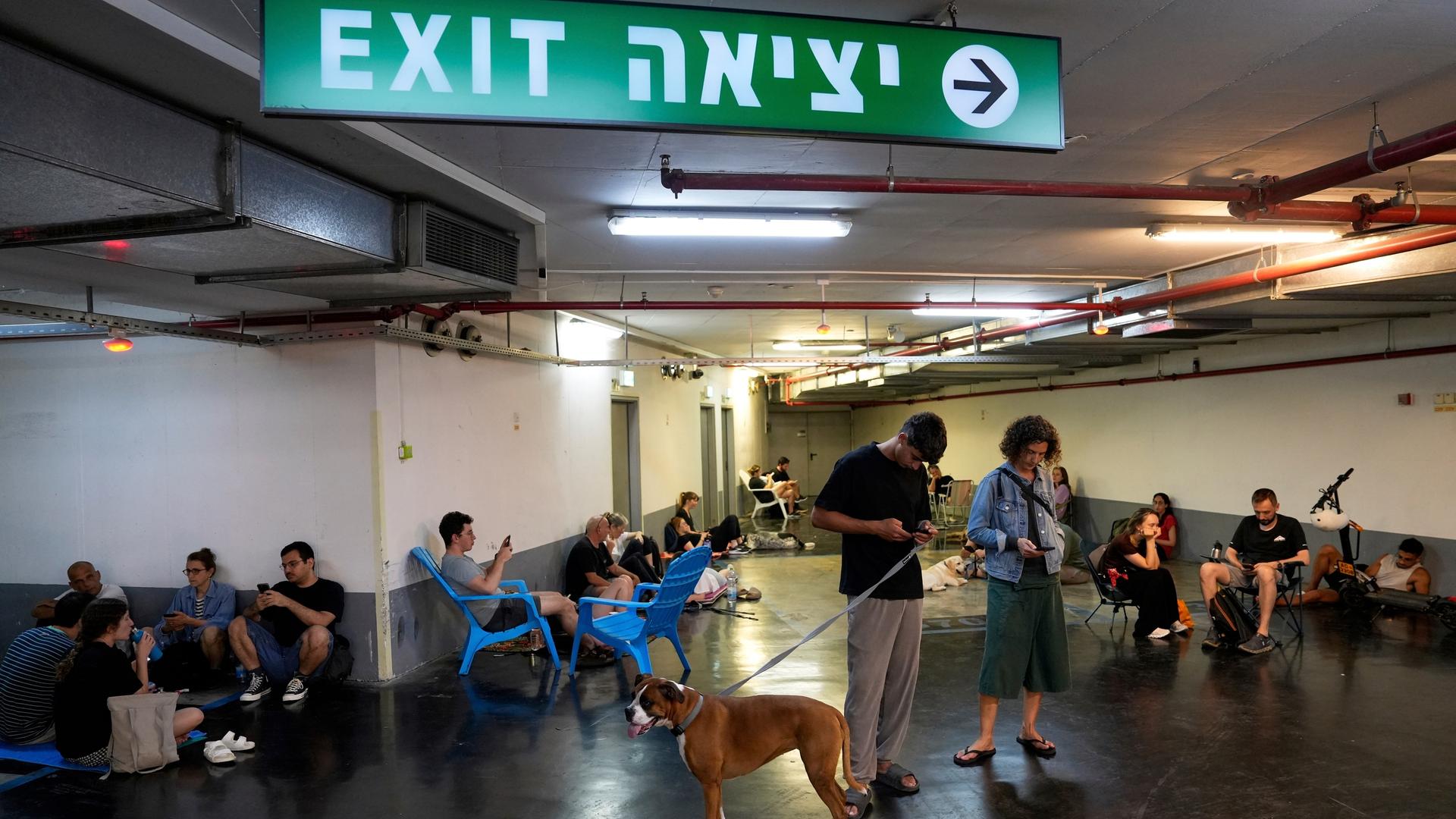 Menschen in einem Schutzraum in Tel Aviv. Etliche schauen auf ihre Mobiltelefone. Ein Paar im Vordergrund hat einen Hund an einer Leine. Menschen in einem Schutzraum in Tel Aviv. Etliche schauen auf ihre Mobiltelefone. Ein Paar im Vordergrund hat einen Hund an einer Leine.
