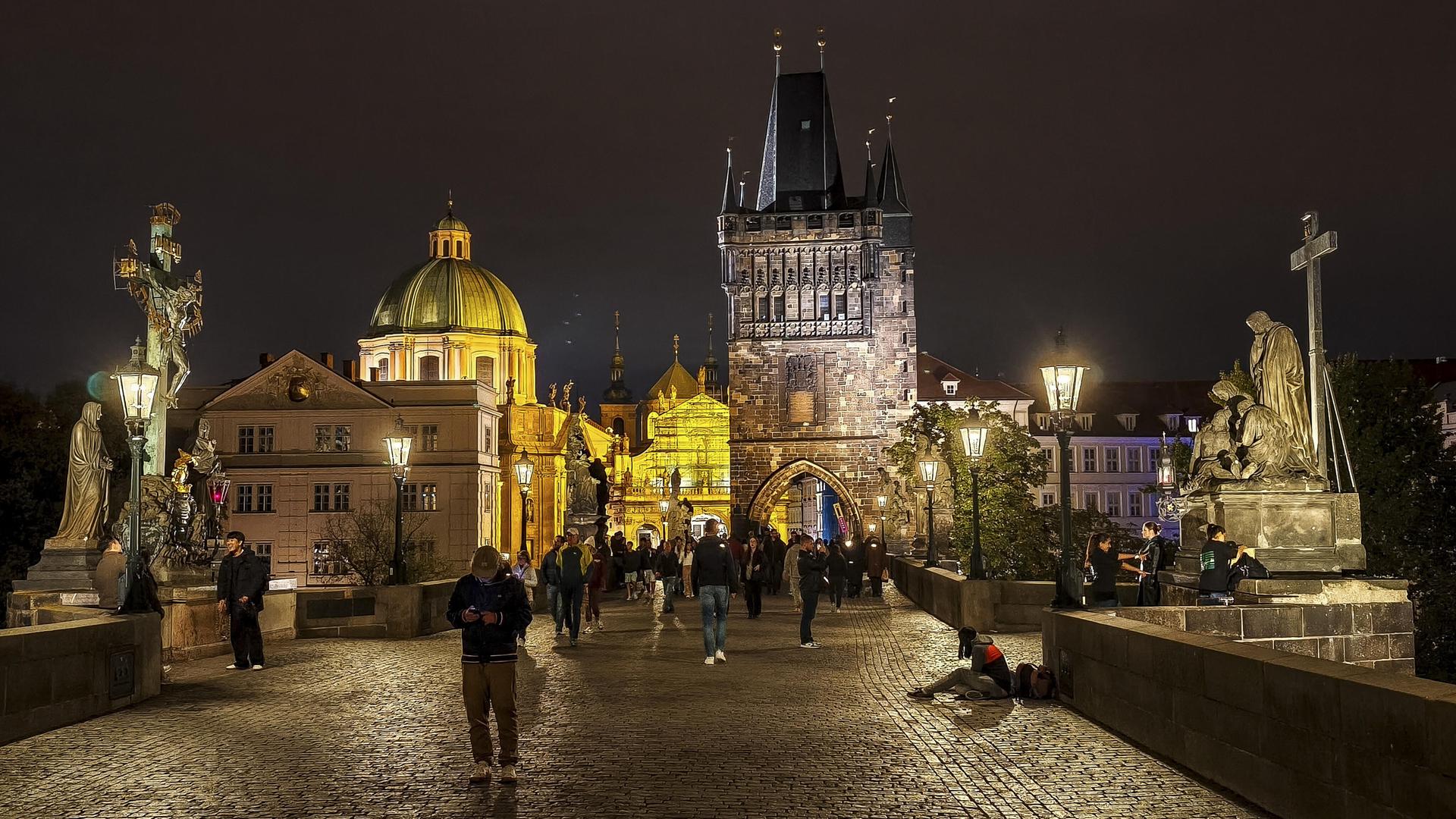 Die Karlsbrücke in Prag bei Nacht, viele Menschen sind unterwegs, einige schauen auf das Display ihres Mobiltelefons.