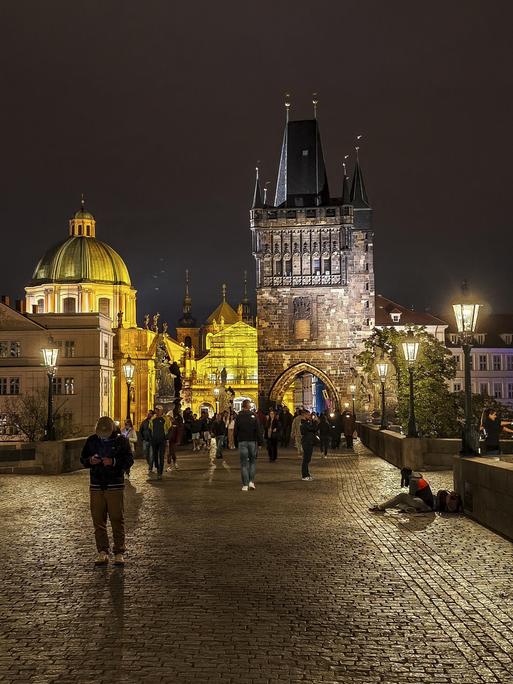 Die Karlsbrücke in Prag bei Nacht, viele Menschen sind unterwegs, einige schauen auf das Display ihres Mobiltelefons.