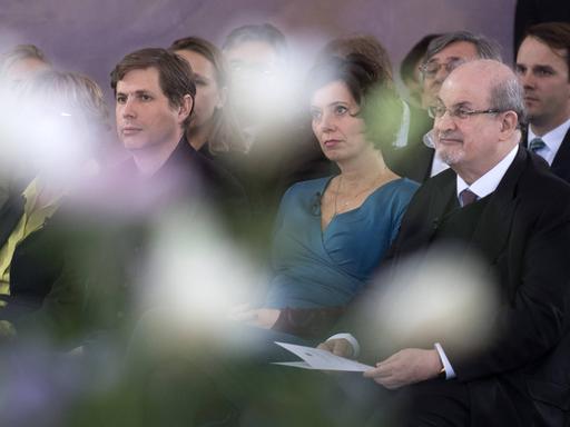 Beim Forum Bellevue zur Zukunft der Demokratie in Berlin saßen am 30. November 2017 die Schriftsteller Daniel Kehlmann, Eva Menasse und Sir Salman Rushdie nebeneinander. Unschärfen verleihen dem Foto Tiefe. Menasse trägt ein türkisblaues Kleid, Rushdie einen dunklen Anzug und Krawatte.