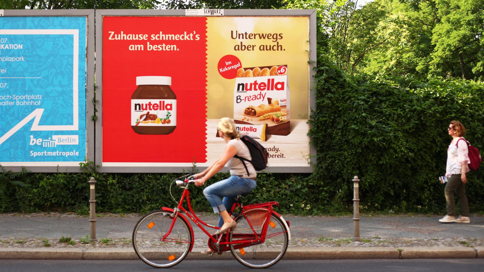 Eine Frau fährt mit dem Fahrrad an einem Plakat mit Webung für Nutella und Nutella B-ready von Ferrero vorbei.