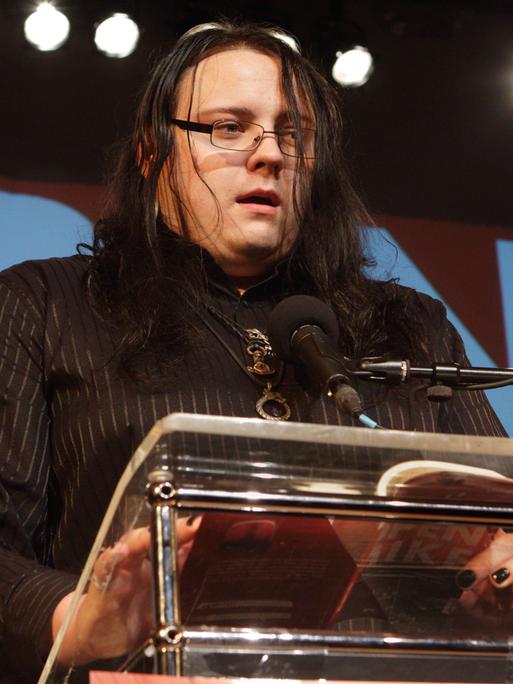Autor Martin Piekar, gekleidet in schwarz, steht an einem Pult und liest aus seinem Preisträgertext. Er trägt seine mittellangen, schwarzen Haare offen, ein schwarzes Brillengestell, einige Ketten um den Hals und hat seine Fingernägel schwarz lackiert. 