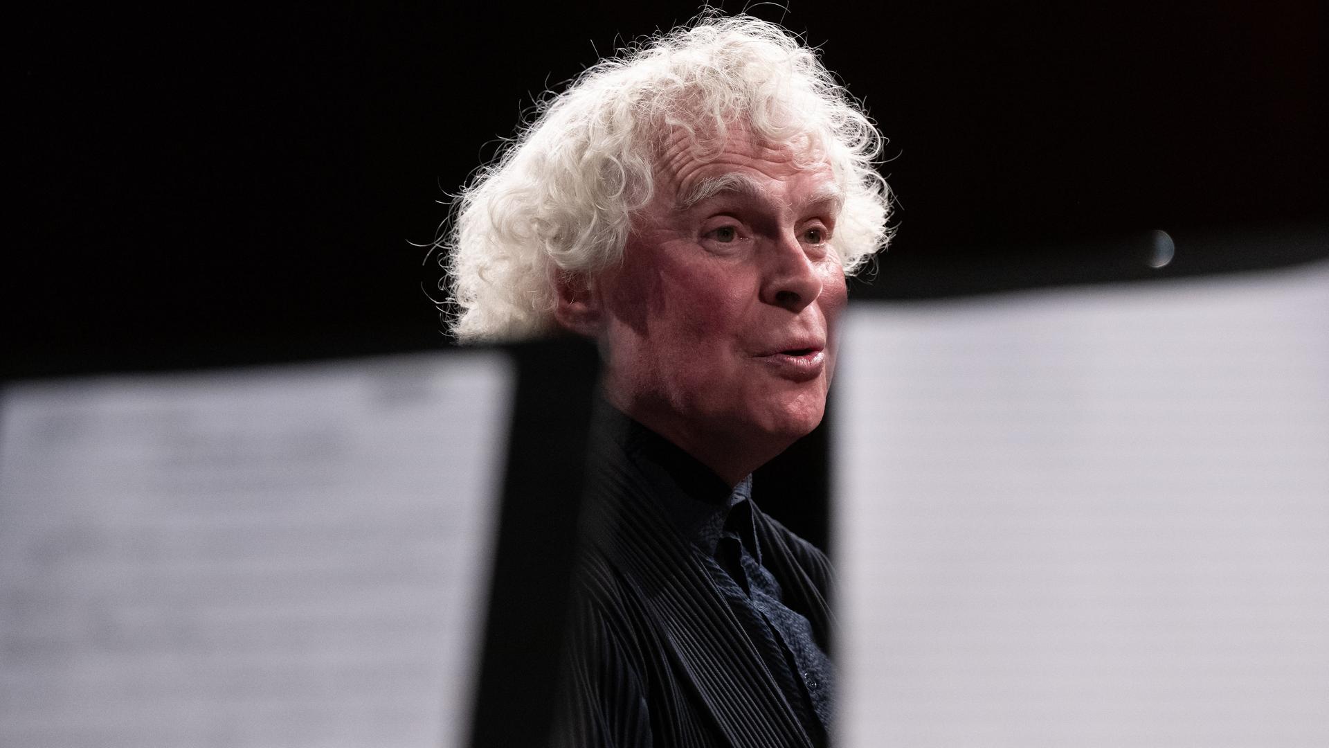 Blick durch Notenpulte hindurch, hinter denen Sir Simon Rattle steht.