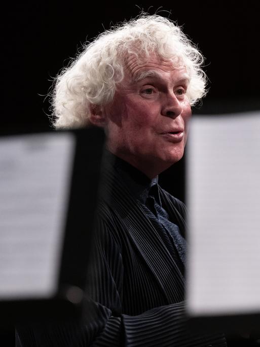 Blick durch Notenpulte hindurch, hinter denen Sir Simon Rattle steht.