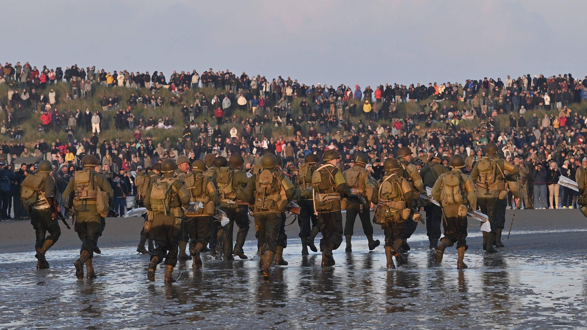 Soldaten gehen an einem Strand entlang, im Hintergrund sitzen und stehen zahlreiche Zuschauer auf einem Hügel.