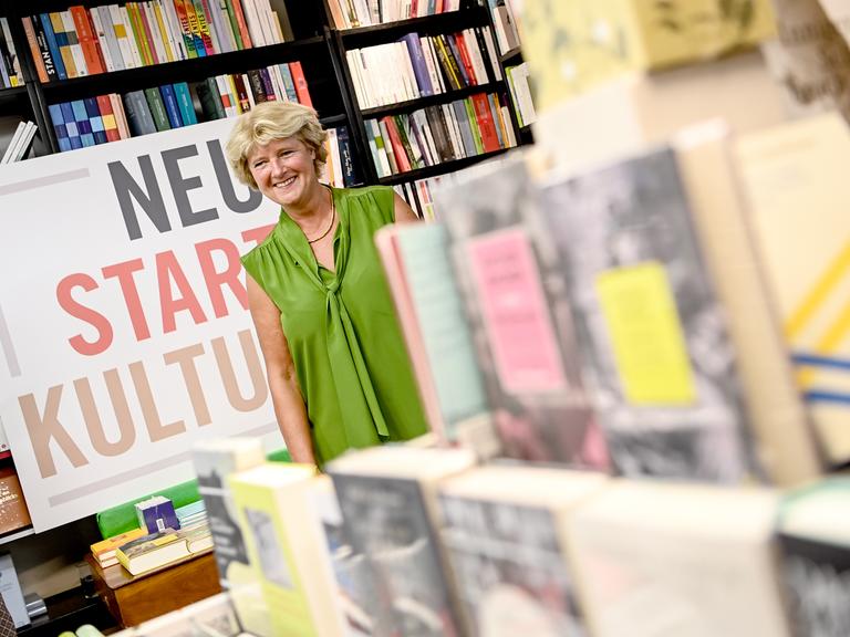 Kulturstaatsministerin Monika Grütters steht inmitten vieler Bücher in einer Buchhandlung, neben ihr ein Schild mit der Aufschrift "Neustart Kultur".