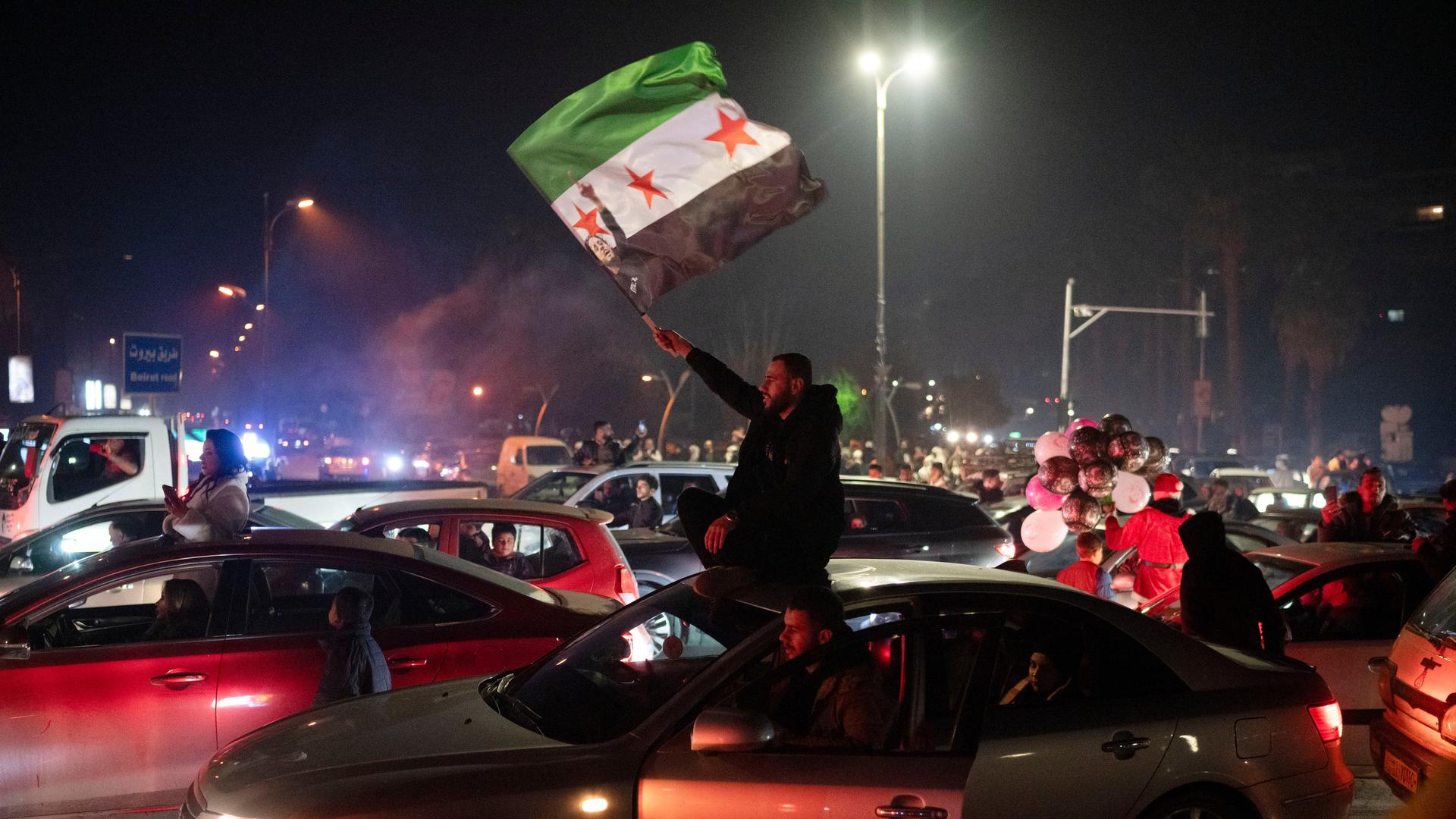 Das Foto zeigt Menschen in der Silvesternacht auf dem Umayyad-Platz in Damaskus. Einer schwenkt eine Fahne der Demokratiebewegung von 2011.