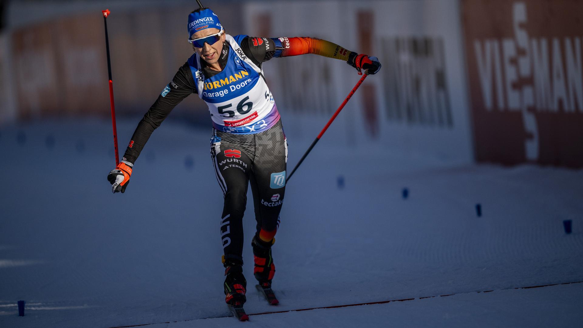 Franziska Preuß überquert die Ziellinie. Sie trägt die typische Sport-Kleidung einer Biathletin und hält zwei Ski-Stöcke in ihren Händen.