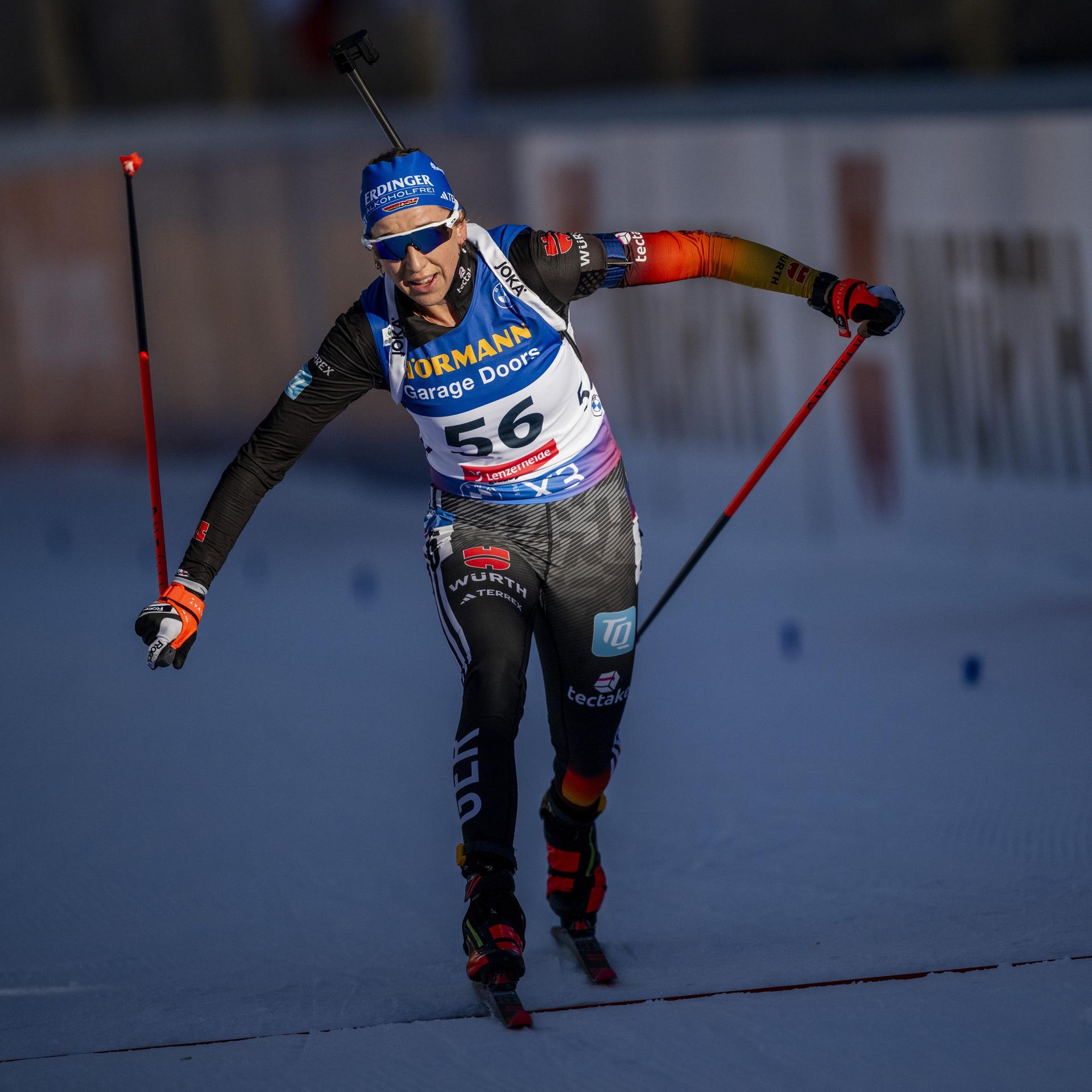 Franziska Preuß überquert die Ziellinie. Sie trägt die typische Sport-Kleidung einer Biathletin und hält zwei Ski-Stöcke in ihren Händen.