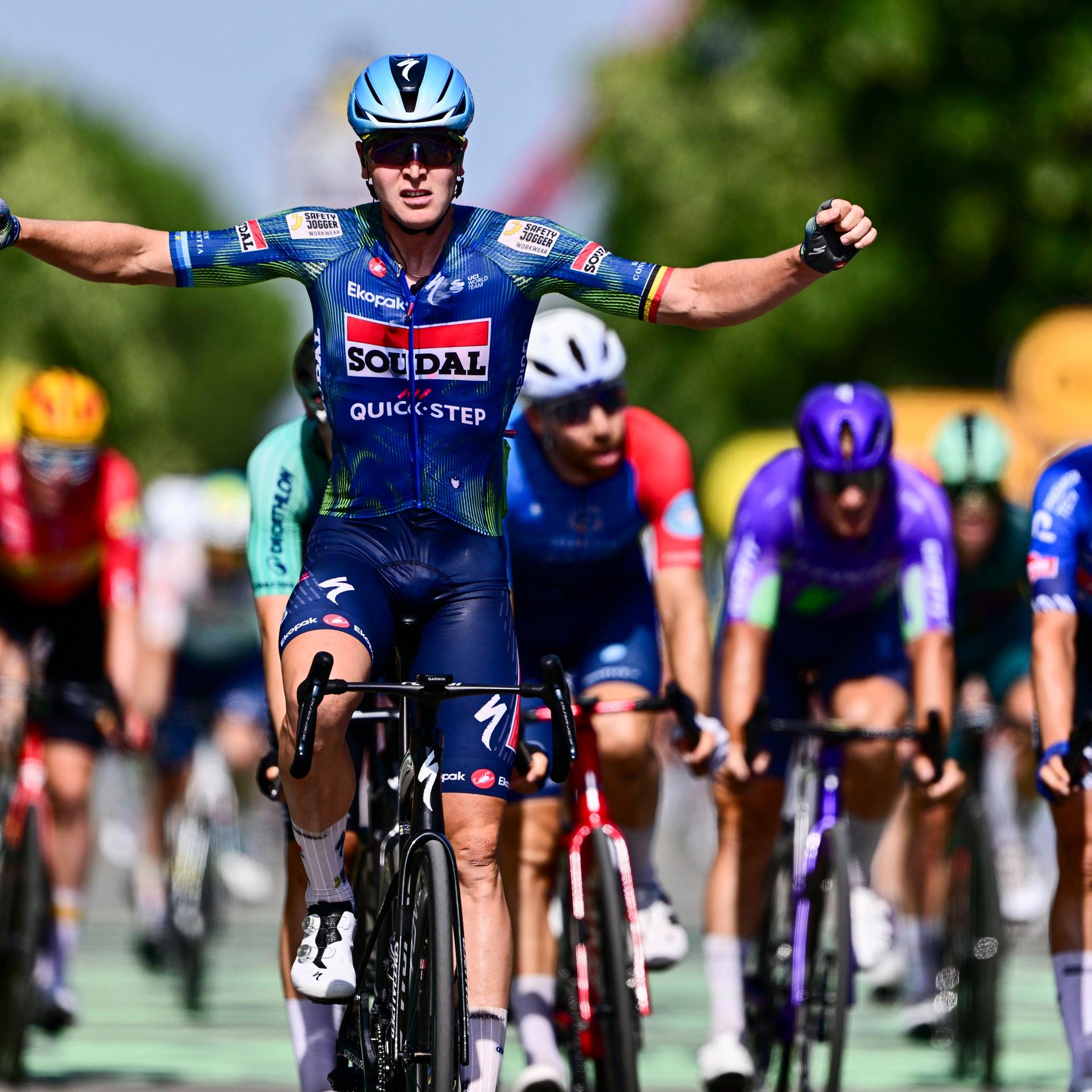 Tour de France: Tim Merlier gewinnt auch die 8. Etappe. Er sitzt auf seinem Rennrad und streckt die Arme nach oben; hinter ihm sind weitere Radrennfahrer zu sehen.
