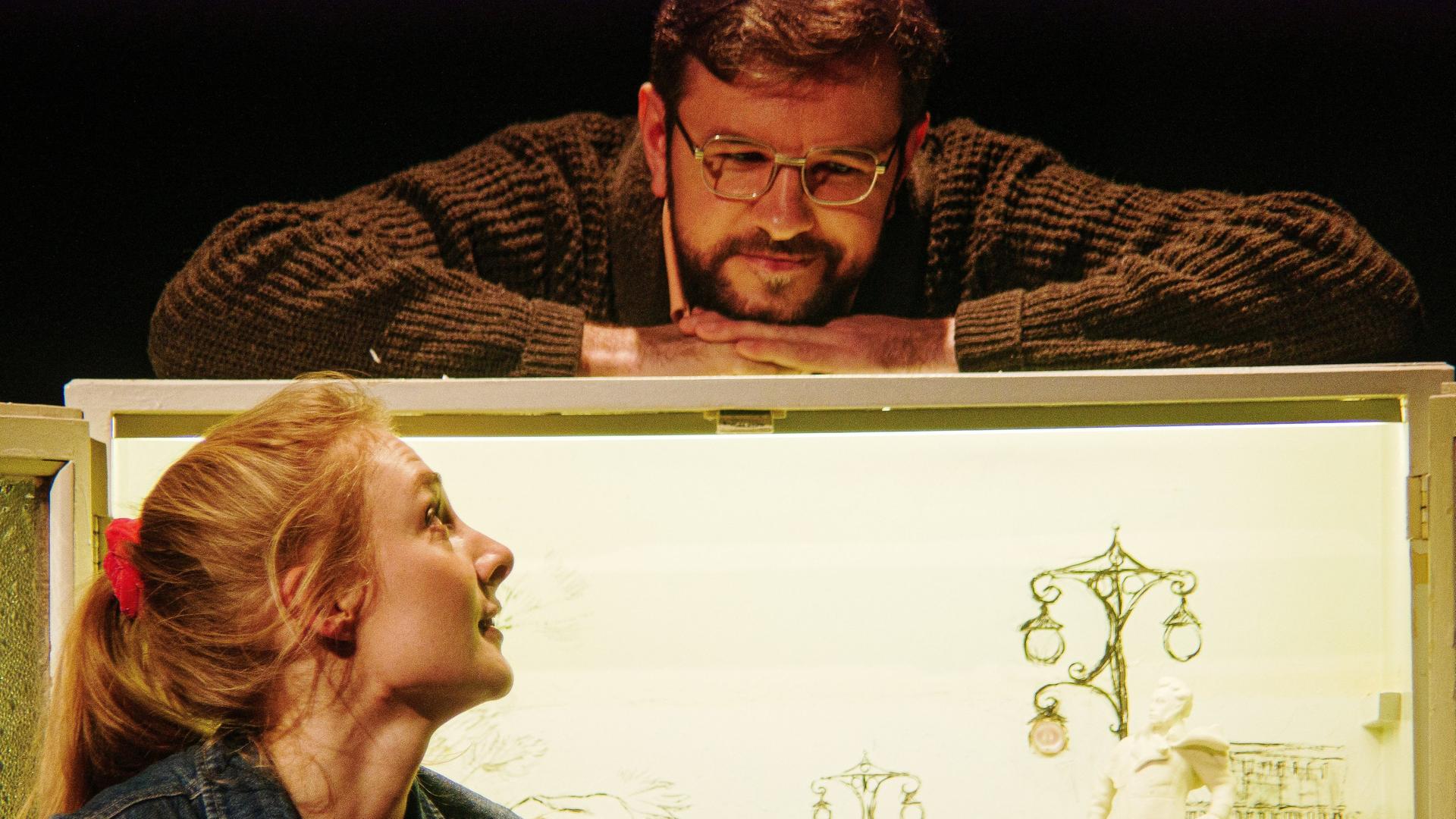 Probeaufnahme des Stücks "Zalot": Eine junge, blonde Frau mit Jeansweste sitzt vor einem Bild. Sie blickt nach oben, wo sich am oberen Rand des Bildes ein Mann befindet. Er trägt eine Brille und einen braunen Pullover, er lächelt.
