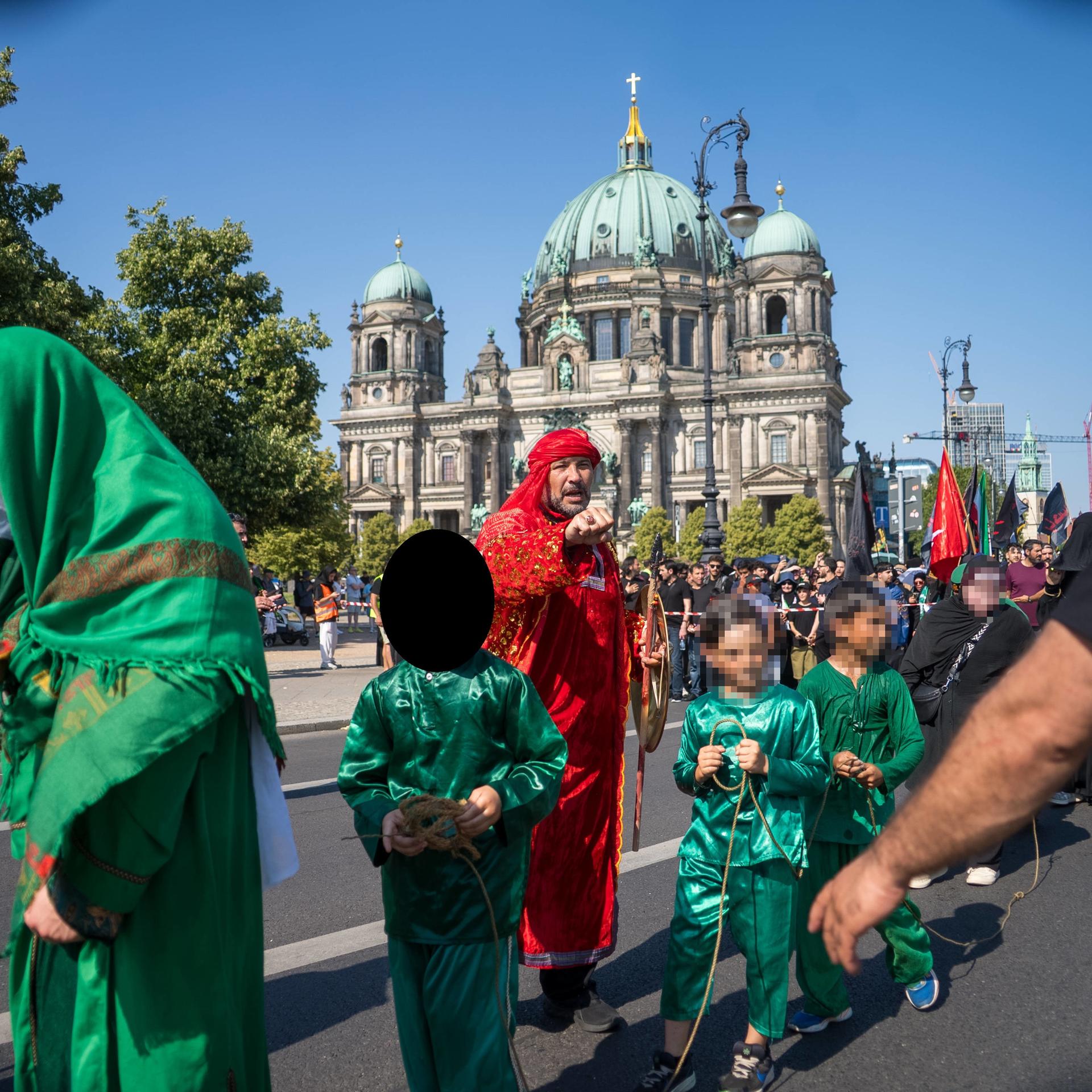 Kinder in langen grünen Gewändern werden an einem Seil über die Straße am Berlin Dom geführt. 
