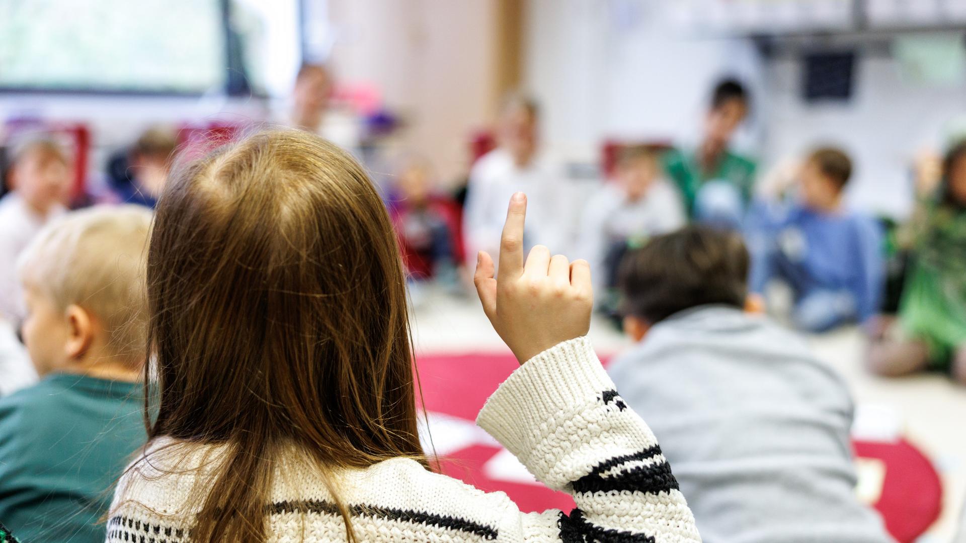 Schüler einer Grundschule sitzen im Sitzkreis auf dem Boden. Ein Mädchen, das von hinten zu sehen ist, meldet sich und hebt den Finger in die Höhe. Es wirkt entspannt.