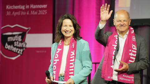 Bundeskanzler Olaf Scholz (SPD) grüßt vom Podium mit erhobener Hand das Publikum auf dem 39. Deutschen Evangelischen Kirchentag in Hannover, links von ihm steht lachend Anja Siegesmund, die Präsidentin des Deutschen Evangelischer Kirchentags. 