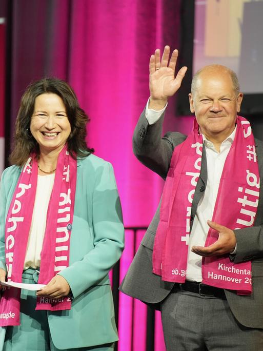 Bundeskanzler Olaf Scholz (SPD) grüßt vom Podium mit erhobener Hand das Publikum auf dem 39. Deutschen Evangelischen Kirchentag in Hannover, links von ihm steht lachend Anja Siegesmund, die Präsidentin des Deutschen Evangelischer Kirchentags. 