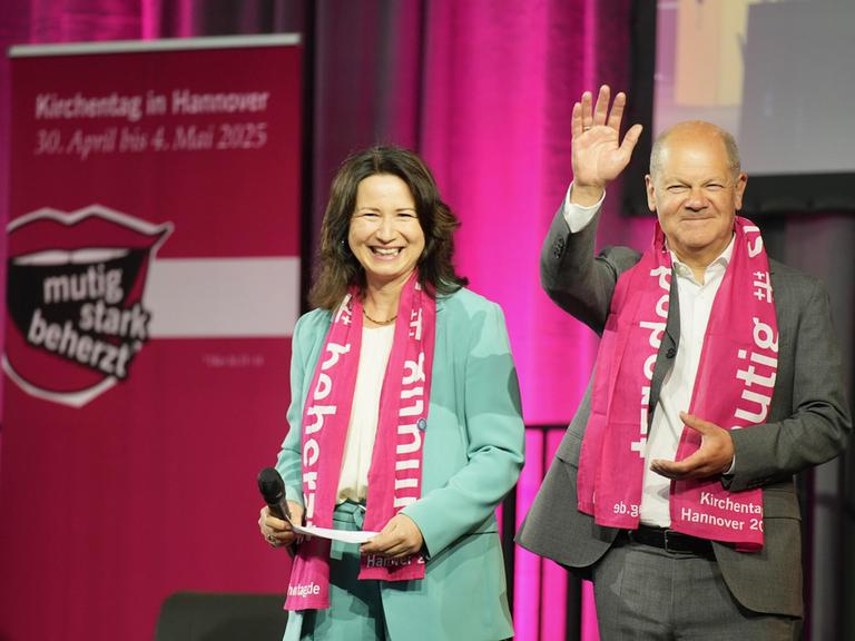 Bundeskanzler Olaf Scholz (SPD) grüßt vom Podium mit erhobener Hand das Publikum auf dem 39. Deutschen Evangelischen Kirchentag in Hannover, links von ihm steht lachend Anja Siegesmund, die Präsidentin des Deutschen Evangelischer Kirchentags. 