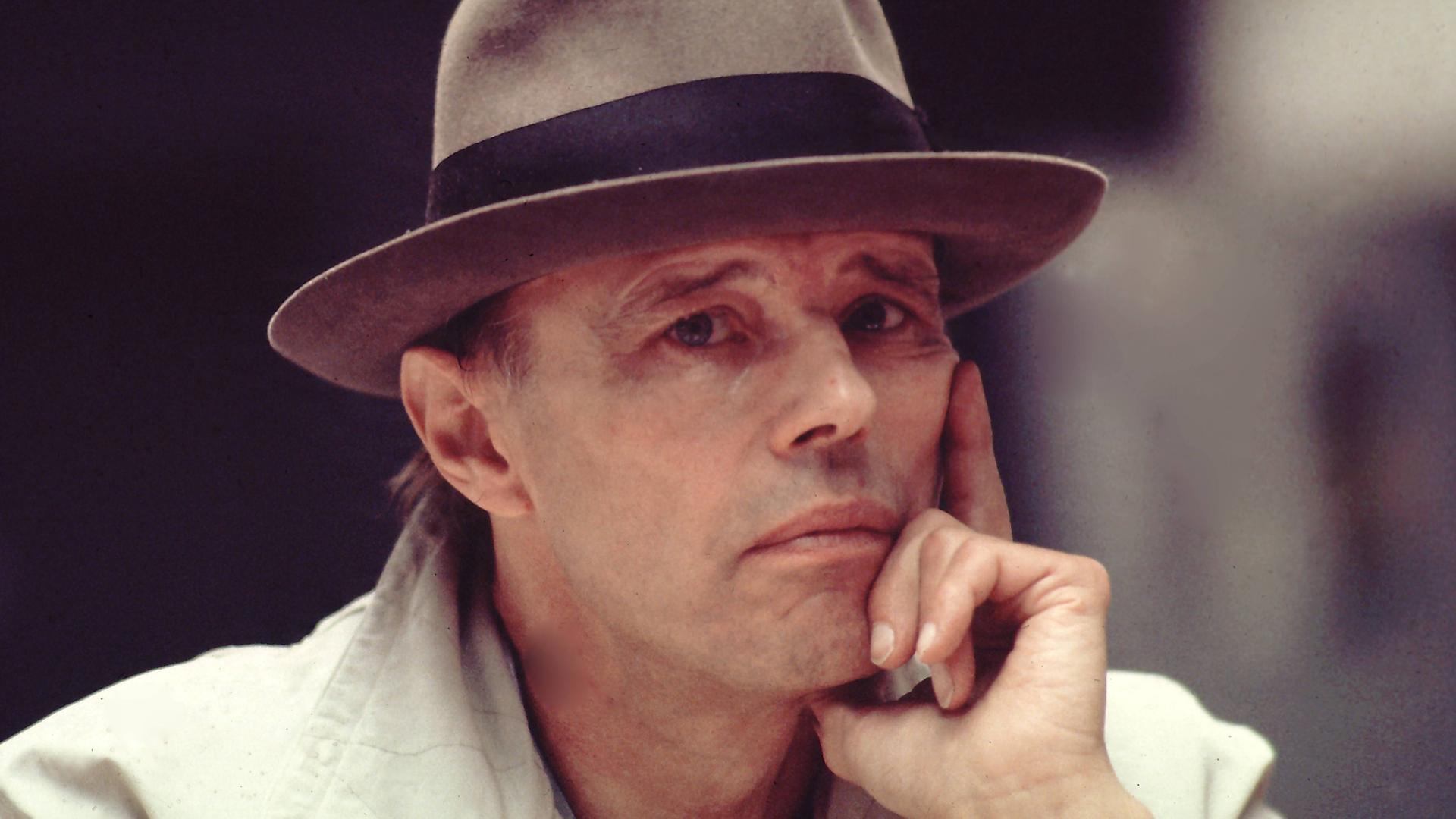 Das Foto zeigt Joseph Beuys von den Schultern aufwärts. Er trägt einen hellgrauen Mantel und einen grauen Hut, um das ein schwarzes Stoffband läuft. Er stützt sein Gesicht auf seine linke Hand und wirkt nachdenklich.