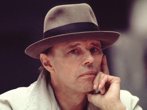 Das Foto zeigt Joseph Beuys von den Schultern aufwärts. Er trägt einen hellgrauen Mantel und einen grauen Hut, um das ein schwarzes Stoffband läuft. Er stützt sein Gesicht auf seine linke Hand und wirkt nachdenklich. Das Foto zeigt Joseph Beuys von den Schultern aufwärts. Er trägt einen hellgrauen Mantel und einen grauen Hut, um das ein schwarzes Stoffband läuft. Er stützt sein Gesicht auf seine linke Hand und wirkt nachdenklich.