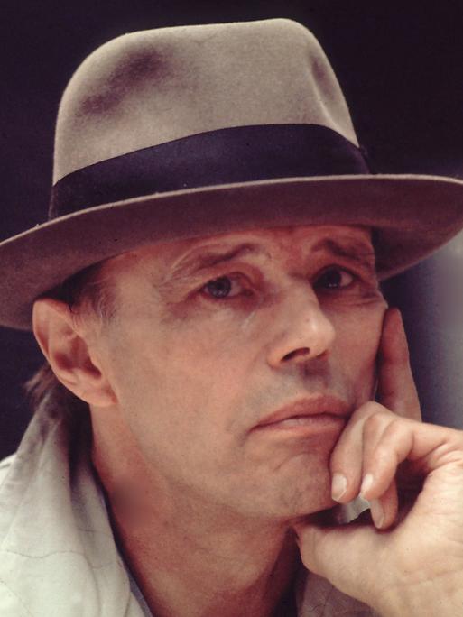 Das Foto zeigt Joseph Beuys von den Schultern aufwärts. Er trägt einen hellgrauen Mantel und einen grauen Hut, um das ein schwarzes Stoffband läuft. Er stützt sein Gesicht auf seine linke Hand und wirkt nachdenklich.