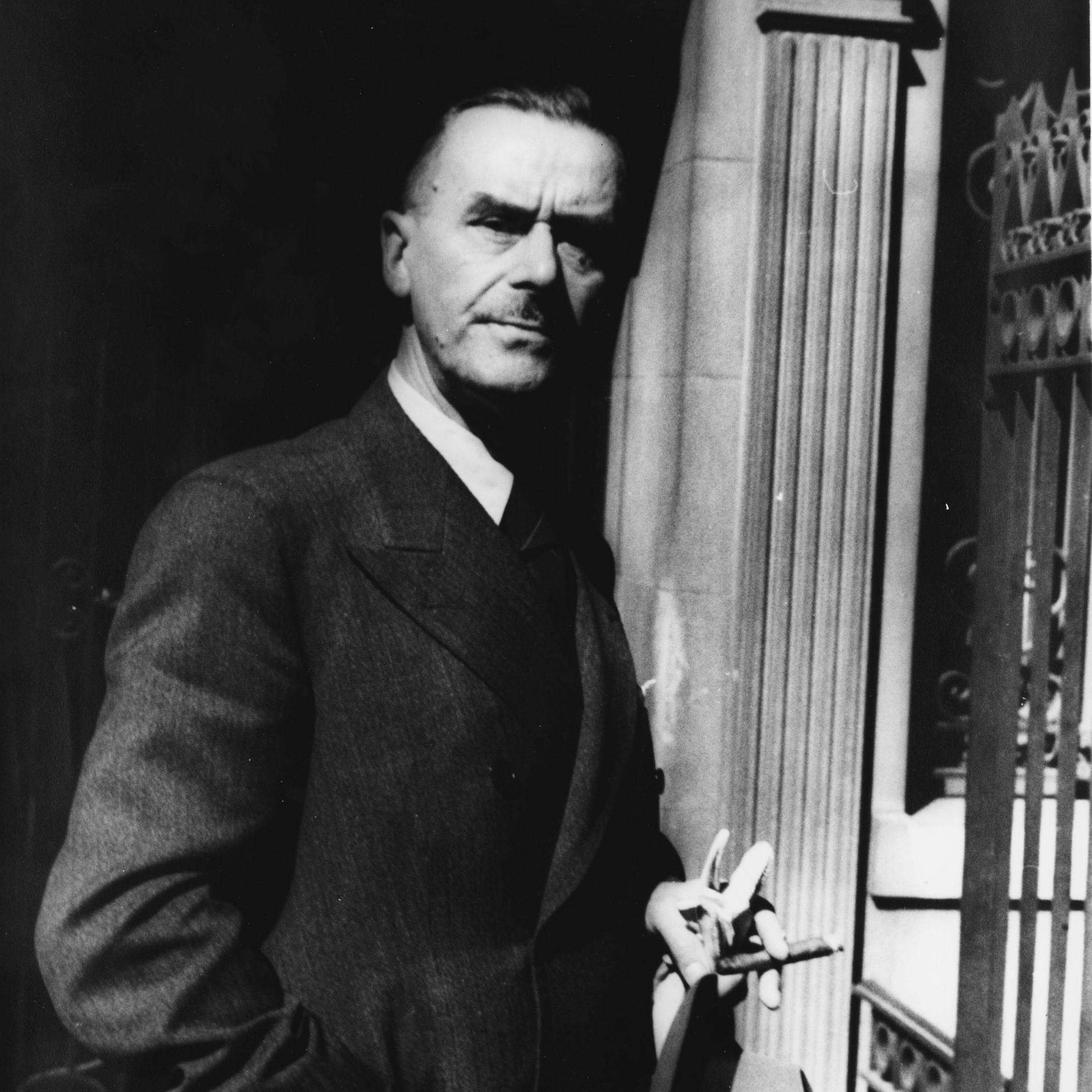 Thomas Mann steht vor einer Säule, hält eine Zigarre und seinen Hut in der Hand und schaut ernst in die Kamera.