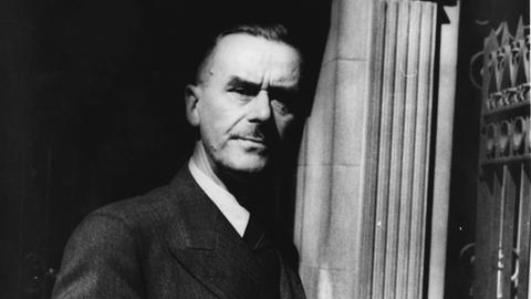 Thomas Mann steht vor einer Säule, hält eine Zigarre und seinen Hut in der Hand und schaut ernst in die Kamera.