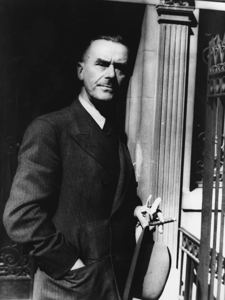 Thomas Mann steht vor einer Säule, hält eine Zigarre und seinen Hut in der Hand und schaut ernst in die Kamera.