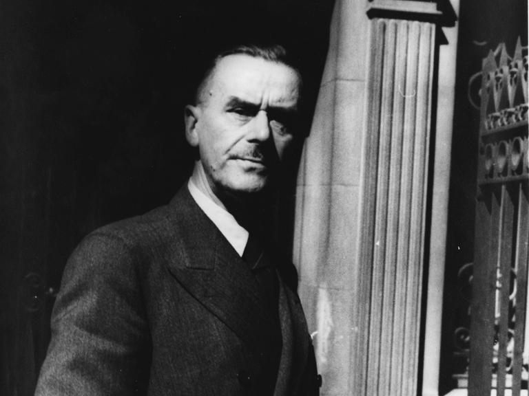Thomas Mann steht vor einer Säule, hält eine Zigarre und seinen Hut in der Hand und schaut ernst in die Kamera.