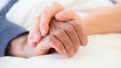 Eine jüngere Hand hält die ältere eines Menschen in einem Bett.