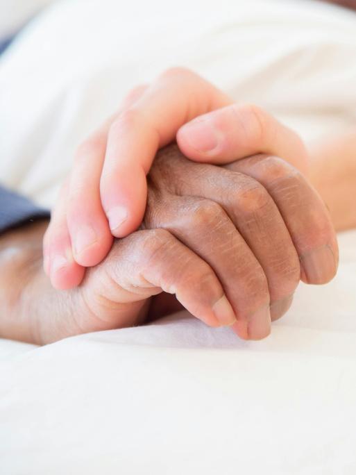 Eine jüngere Hand hält die ältere eines Menschen in einem Bett.