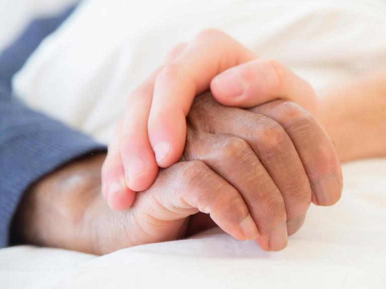 Eine jüngere Hand hält die ältere eines Menschen in einem Bett.