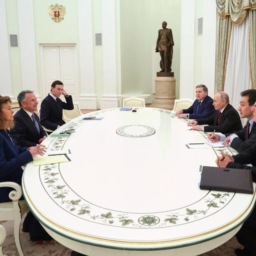 Das Bild zeigt zwei Verhandlungs-Delegationen an einem großen und ovalen weißen Tisch. Sie sitzen sich an den langen Enden gegenüber und lächeln sich freundlich an.