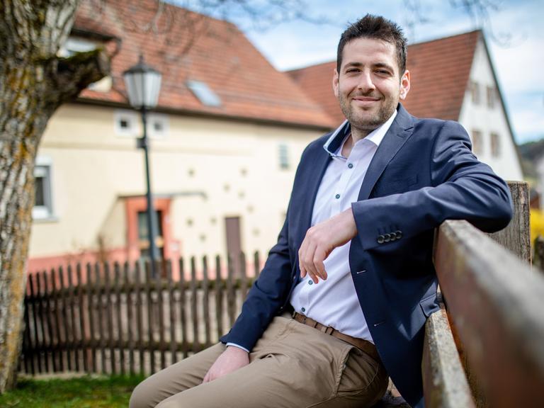 Ryyan Alshebl sitzt im Anzug in Ostelsheim auf einer Parkbank und lächelt in die Kamera.