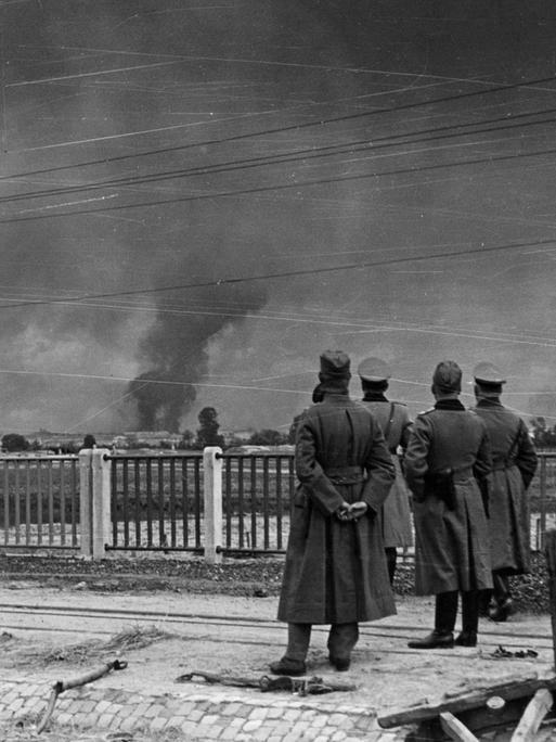 Im Vordergrund stehen deutsche Soldaten der Wehrmacht und beobachten die Angriffe auf Warschau.