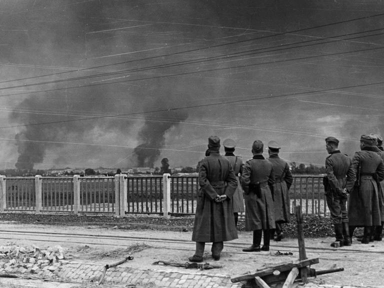 Im Vordergrund stehen deutsche Soldaten der Wehrmacht und beobachten die Angriffe auf Warschau.
