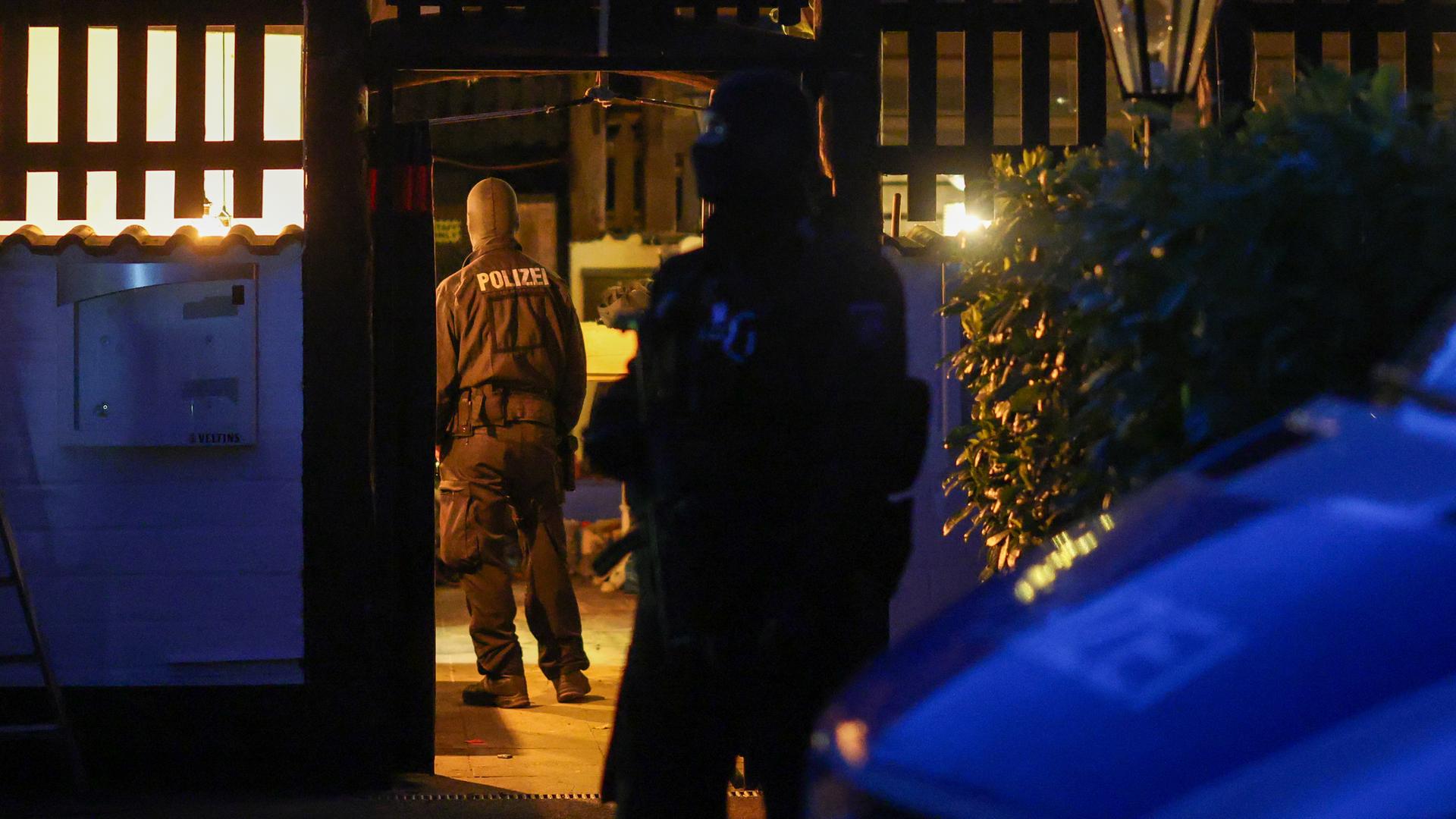 Man sieht zwei Polizisten von hinten, die während einer Razzia den Eingang zu einem beleuchteten Geländer der Hells Angels betreten.