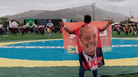 Eine Mann hält ein großes Porträt des brasilianischen Präsidenten hoch, während er neben einer riesigen brasilianischen Flagge auf einer einer Wahlkampfkundgebung in Rio de Janeiro steht. Lula ist auf der in der Ferne gelegenen Bühne zu sehen. Eine Mann hält ein großes Porträt des brasilianischen Präsidenten hoch, während er neben einer riesigen brasilianischen Flagge auf einer einer Wahlkampfkundgebung in Rio de Janeiro steht. Lula ist auf der in der Ferne gelegenen Bühne zu sehen.