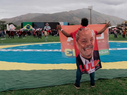 Eine Mann hält ein großes Porträt des brasilianischen Präsidenten hoch, während er neben einer riesigen brasilianischen Flagge auf einer einer Wahlkampfkundgebung in Rio de Janeiro steht. Lula ist auf der in der Ferne gelegenen Bühne zu sehen. Eine Mann hält ein großes Porträt des brasilianischen Präsidenten hoch, während er neben einer riesigen brasilianischen Flagge auf einer einer Wahlkampfkundgebung in Rio de Janeiro steht. Lula ist auf der in der Ferne gelegenen Bühne zu sehen.