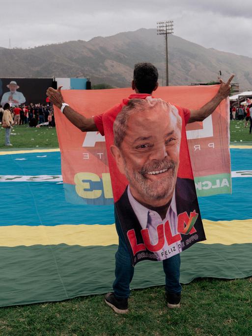 Eine Mann hält ein großes Porträt des brasilianischen Präsidenten hoch, während er neben einer riesigen brasilianischen Flagge auf einer einer Wahlkampfkundgebung in Rio de Janeiro steht. Lula ist auf der in der Ferne gelegenen Bühne zu sehen.