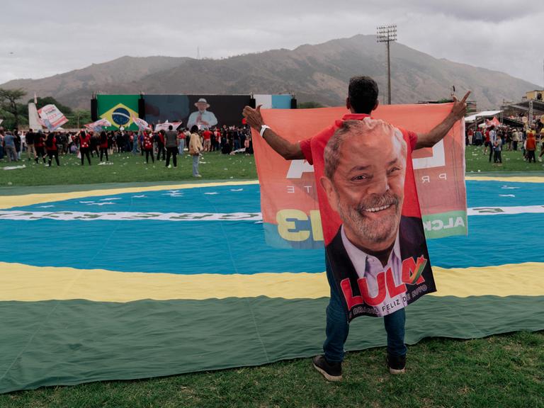 Eine Mann hält ein großes Porträt des brasilianischen Präsidenten hoch, während er neben einer riesigen brasilianischen Flagge auf einer einer Wahlkampfkundgebung in Rio de Janeiro steht. Lula ist auf der in der Ferne gelegenen Bühne zu sehen.