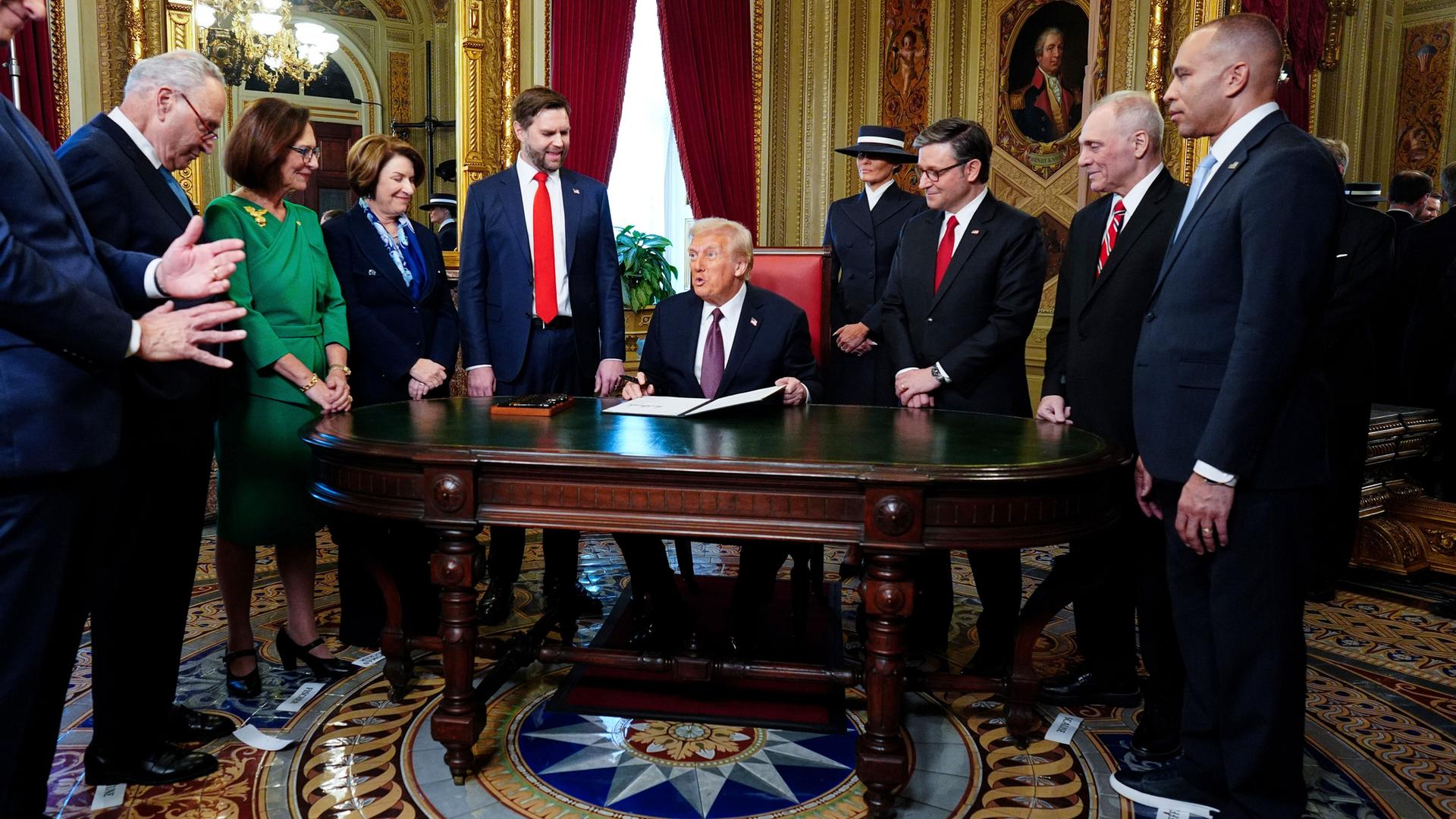 Das Foto zeigt US-Präsident Trump im Kapitol an einem Schreibtisch, umgeben von zahlreichen Menschen.