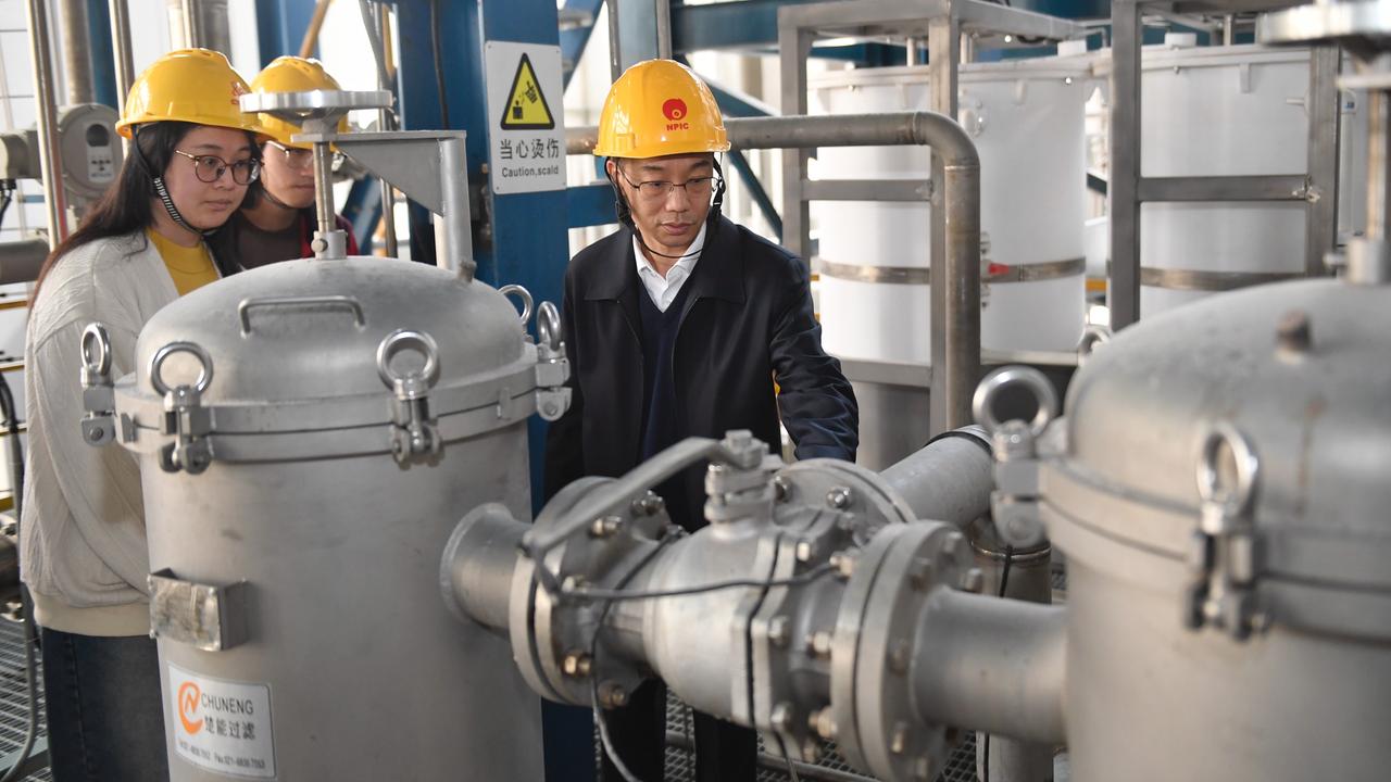 Zwei chinesische Männer und eine Frau tragen gelbe Sicherheitshelme und inspizieren silber-graue Kessel-Konstruktionen und Rohre. 