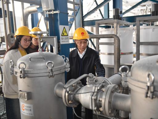 Zwei chinesische Männer und eine Frau tragen gelbe Sicherheitshelme und inspizieren silber-graue Kessel-Konstruktionen und Rohre. 