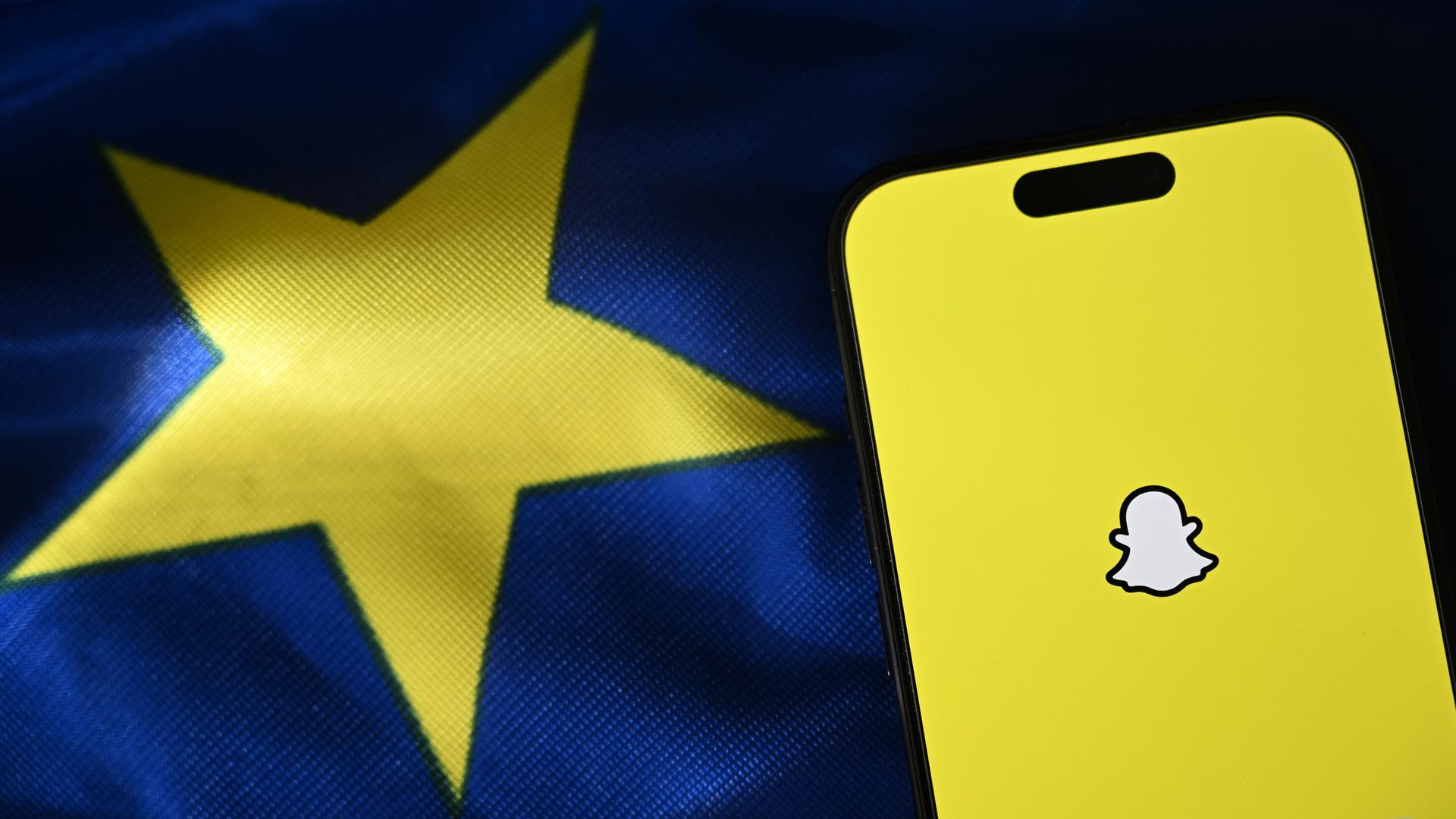 Eine Illustration der App Snapchat auf einem Smartphone, das auf einer EU-Flagge liegt.