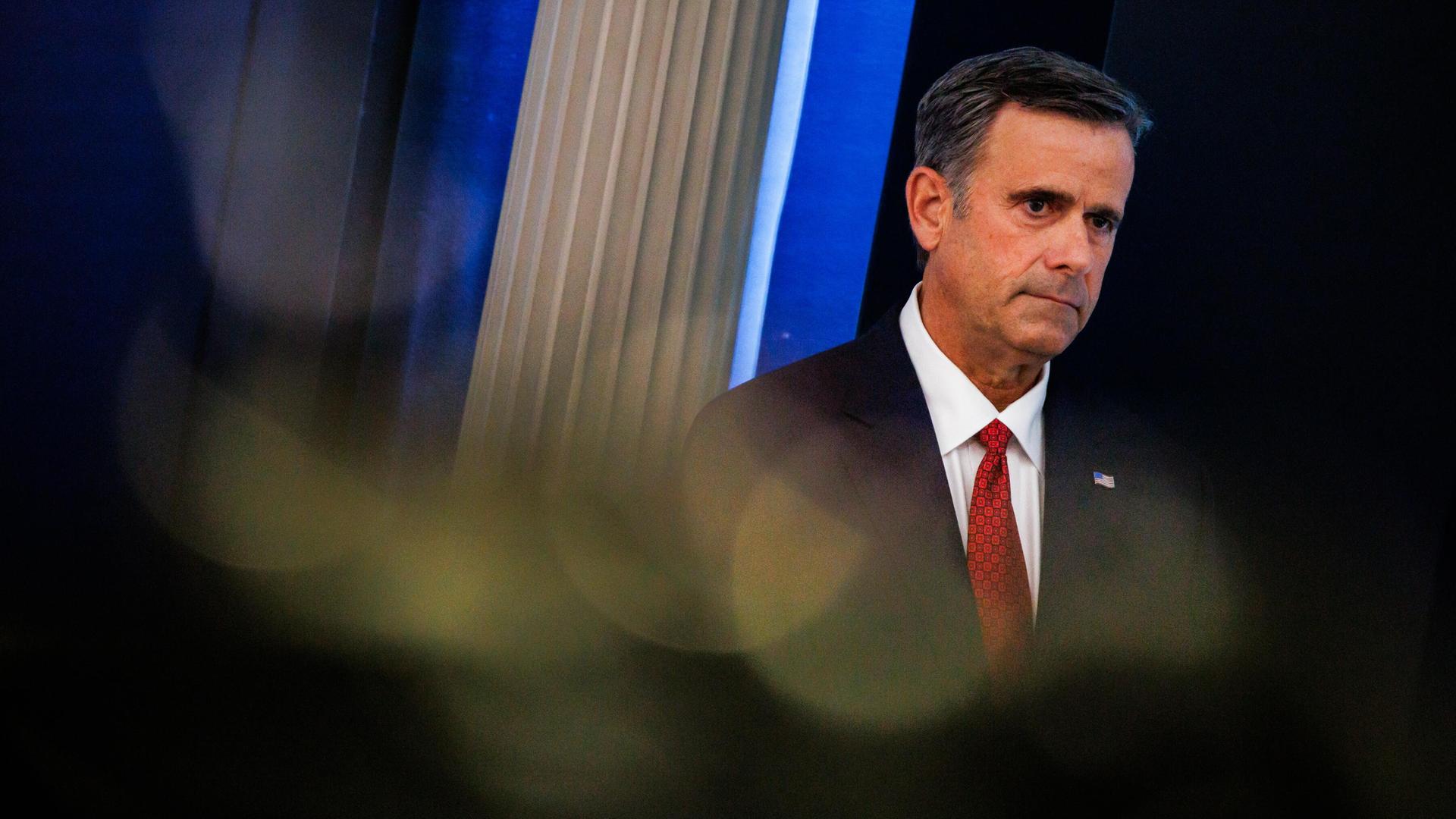 John Ratcliffe trägt eine schwarze Anzugjacke, ein weißes Hemd und eine rote Krawatte und steht vor einem dunklen Hintergrund. Sein Blick geht zur Seite. John Ratcliffe trägt eine schwarze Anzugjacke, ein weißes Hemd und eine rote Krawatte und steht vor einem dunklen Hintergrund. Sein Blick geht zur Seite.