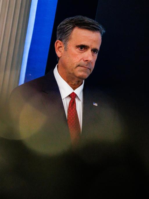 John Ratcliffe trägt eine schwarze Anzugjacke, ein weißes Hemd und eine rote Krawatte und steht vor einem dunklen Hintergrund. Sein Blick geht zur Seite.